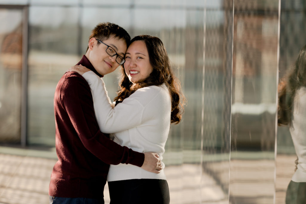 An engaged couple holds each other in an embrace and smiles towards the camera. They are on an outdoor patio with large windows and reflections surround them.