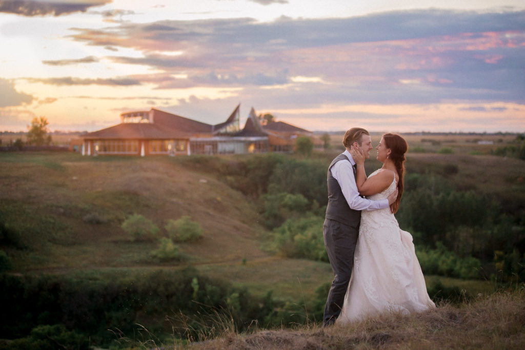 A newly married couple hold each other and make silly faces while they stand at the top of a hill on their wedding day. In the background behind them sits the large outline of the wedding venue they were married in.