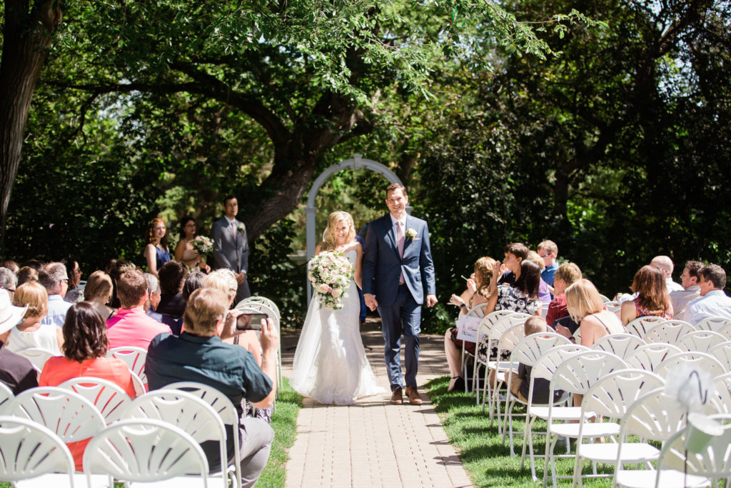 A newly married couple walks up the aisle together at an outdoor ceremony while their friends and family sit in chairs surrounding them. 